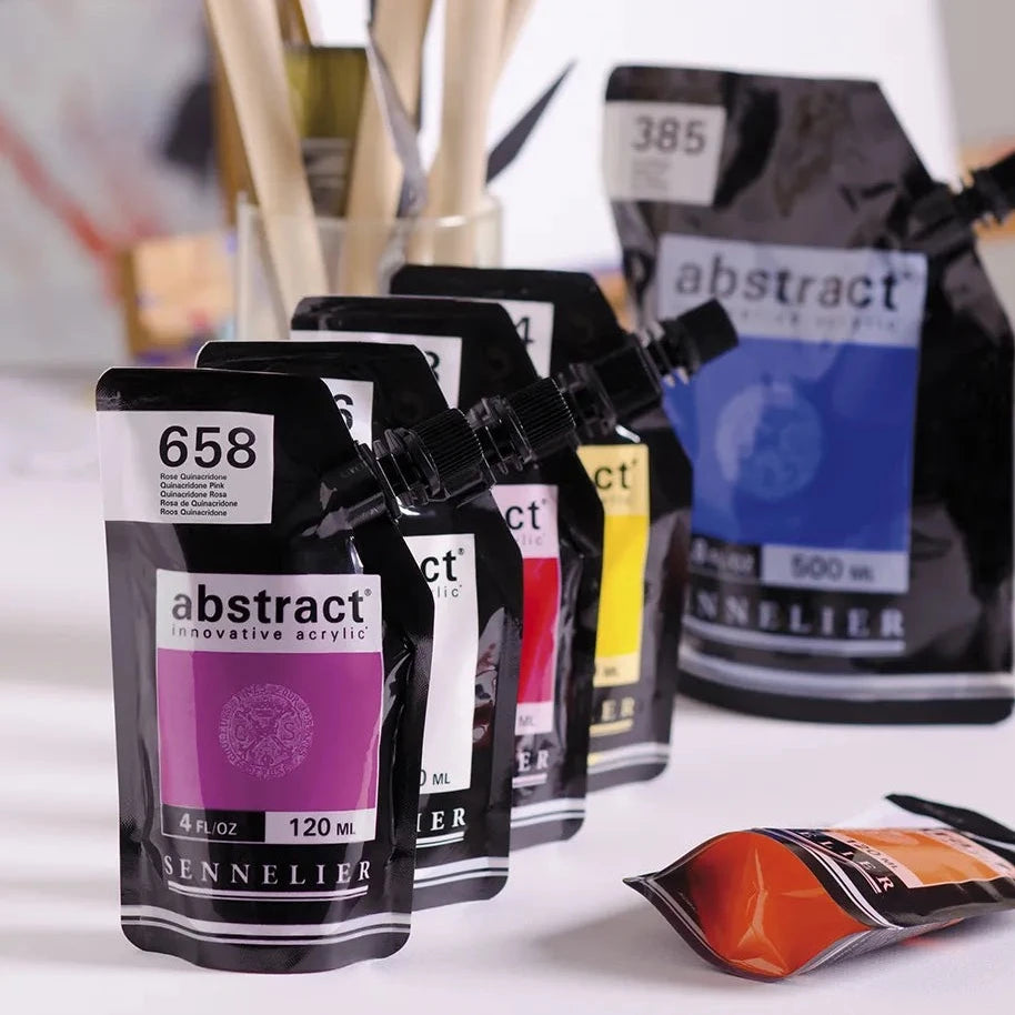 Studio background with paintbrushes in a glass and 4 120ml acrylic packets lined up in front of a 500ml packet and one small pack tipped over on table